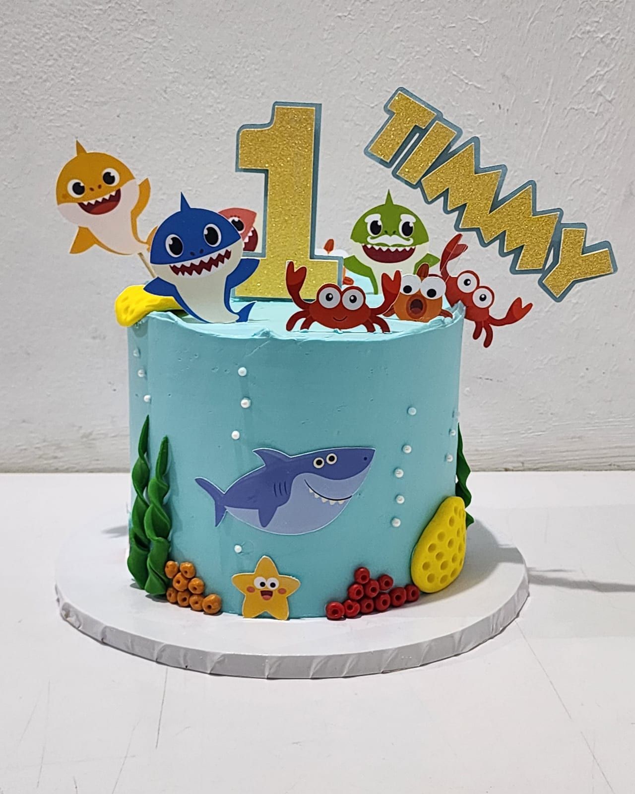 Tall pale blue cake decorated with cartoon sharks, crabs, starfish, seaweed and sugar pearls, with a gold number one topper