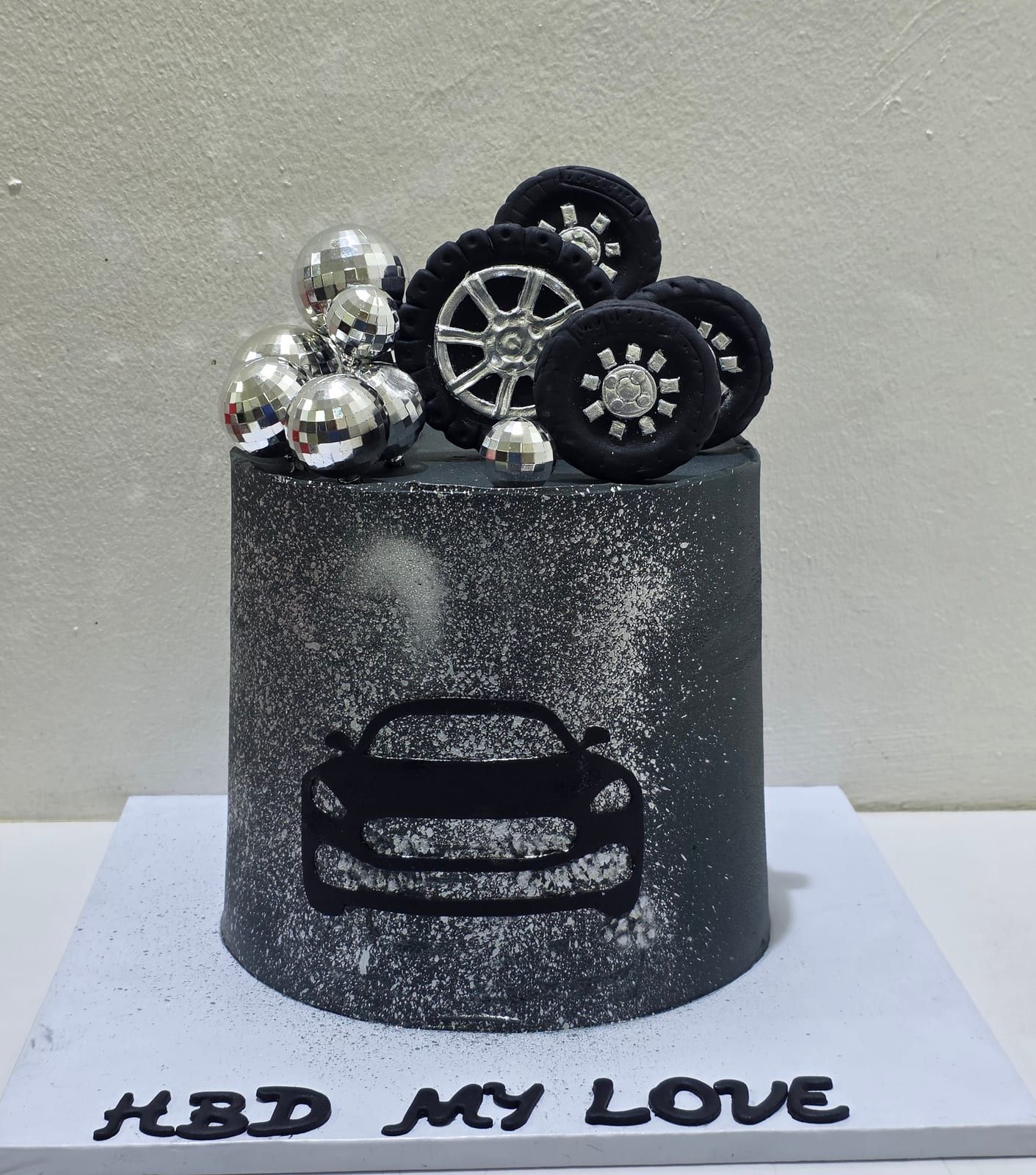 Tall black cake dusted with silver, with a car silhouette on the front, sugar tyres and a wheel rim stacked on top with silver spheres, and "HBD My Love" piped in black at the base