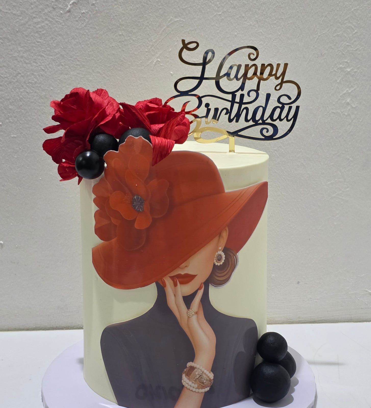 A tall ivory cake with an edible-print portrait of a woman in a wide red hat, crowned with deep red sugar flowers, made by Sunset Cakes