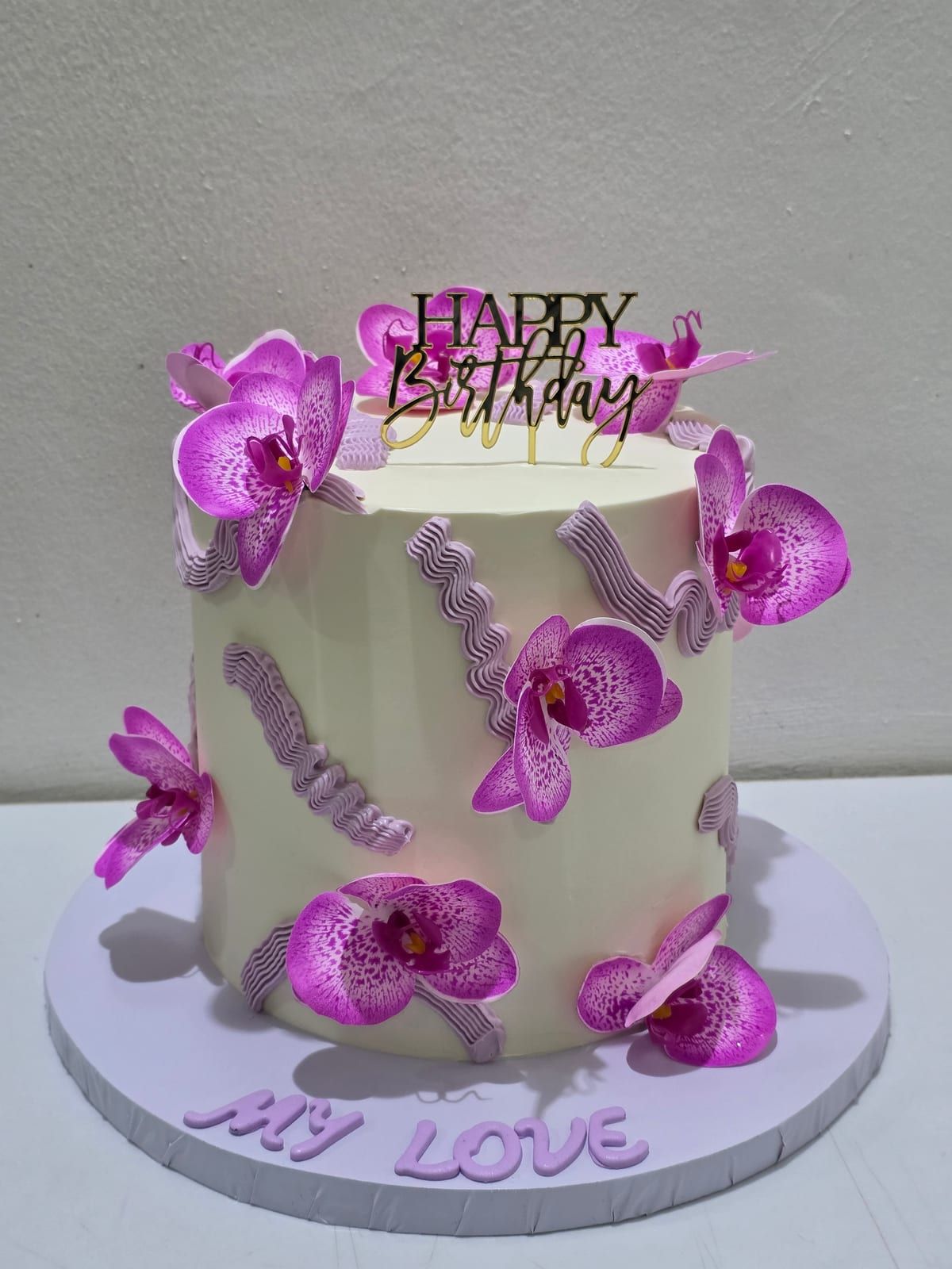 Tall ivory cake covered in magenta sugar orchids and thin purple ribbon curls, with a black "Happy Birthday" topper and "My Love" piped in purple at the base