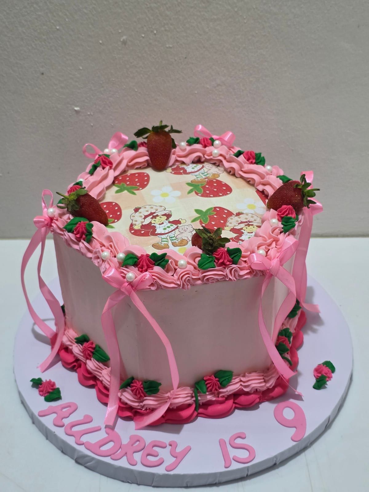Ivory cake with pink vintage-style piping, a printed strawberry picture on top, fresh strawberries around the edge, pink ribbons down the sides and "Audrey is 9" piped in pink at the base
